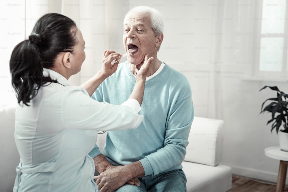 Montrez votre langue. Homme âgé aux cheveux gris et agréable, assis sur le canapé, en train de subir un examen médical et de montrer sa langue.
