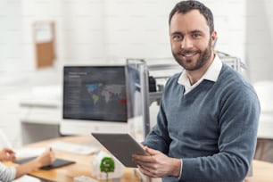 New information. Joyful bristled young man leaning on the desk in the office and posing for the camera while reading from tablet new data regarding his project