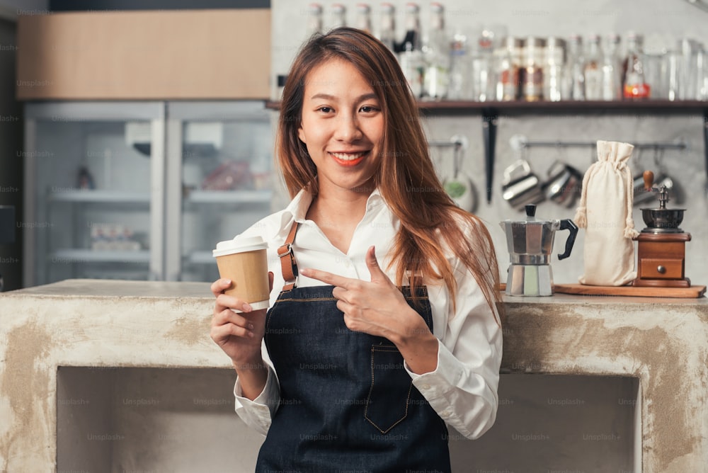 Close up of a young Asian female barista hold a cup of coffee serving to her customer with smile surrounded with bar counter background. Young female barista and her small shop. Food and drink concept