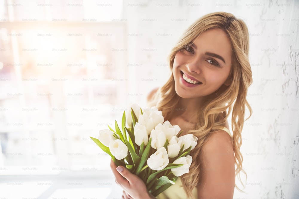 Portrait of attractive young woman with tulips is standing in light room and smiling. Happy international women's day! Celebrating 8th of March.
