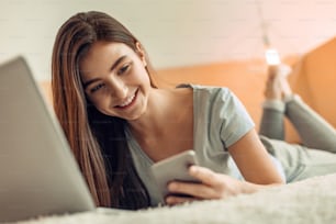 필요한 일시 중지. 유쾌한 쾌활한 십대 소녀가 침대에 누워 휴대폰으로 메시지를 읽고, 밝게 웃으며, 공부를 잠시 멈췄습니다.