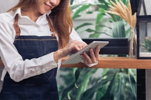 Young Asian female barista using electronic tablet in her own coffee shop while takeing break in warm afternoon. Young female barista and her modern small coffee shop. Food and drink industry concept.