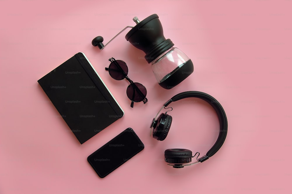 Stilvolle schwarze Sonnenbrille, Smartphone, Kopfhörer, Kaffeemühle und Notizbuch auf rosa Hintergrund, flach legen. Modernes Hipster-Image. schwarze Gegenstände auf rosa Papier. Instagram-Bloggen
