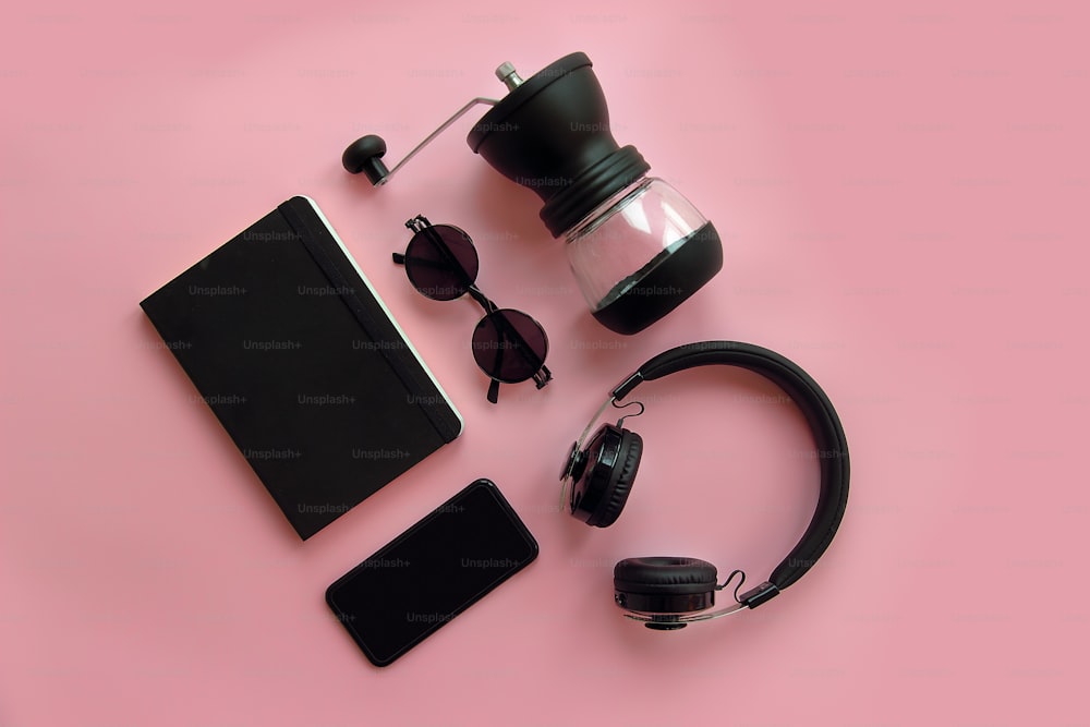 Elegantes gafas de sol negras, teléfono inteligente, auriculares, molinillo de café y cuaderno sobre fondo rosa, plano. Imagen hipster moderna. Artículos negros sobre papel rosa. Blogs de Instagram