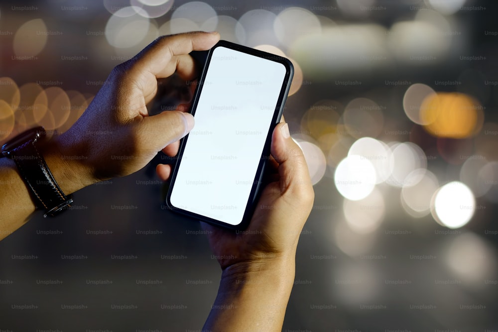 Primer plano de manos masculinas que muestran un teléfono inteligente con pantalla en blanco en el exterior, luces bokeh