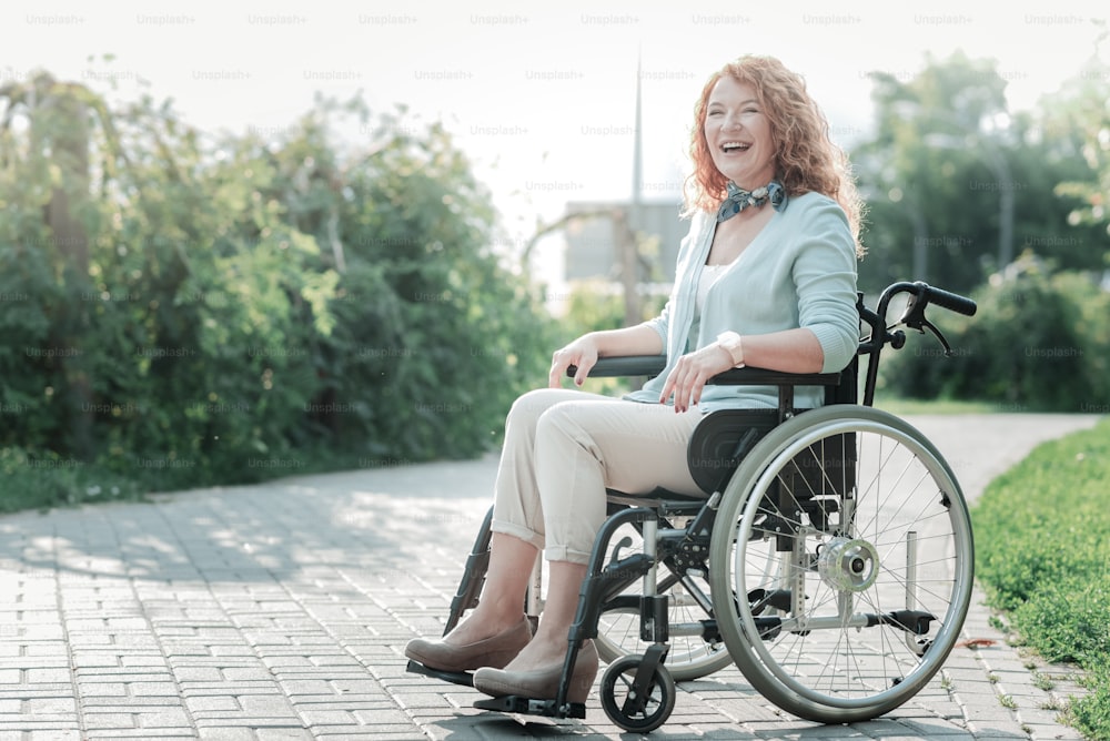 Feeling happiness. Attractive female person keeping smile on her face and sitting on her wheelchair while being outdoors