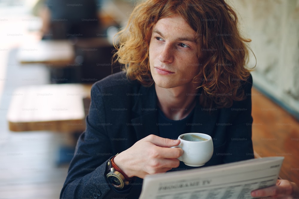 Nahaufnahme-Porträt. Junger interessanter Mann mit lockigen rötlichen Haaren, trägt Jacke und Vintage-Uhr, liest Zeitung und trinkt Kaffee, während er an einem Holztisch in einem Café im Freien sitzt.