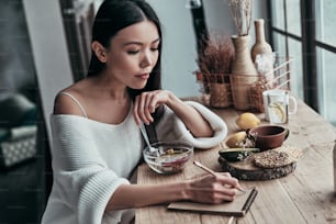 Attractive young woman eating healthy breakfast and writing something down while sitting near the window at home