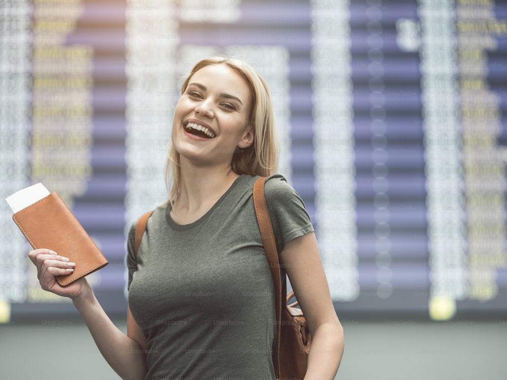 Portrait of laughing female keeping tickets in arm while standing opposite timetable. Glad tourist concept