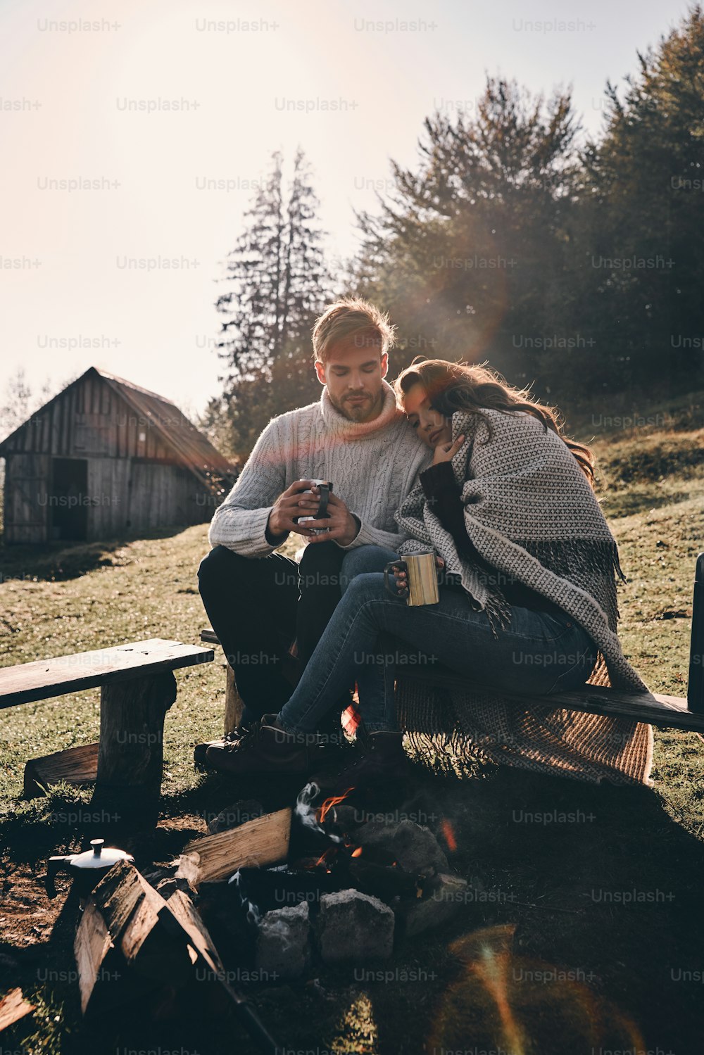 山のキャンプファイヤーのそばに座りながら朝のコーヒーを飲む美しい若いカップル