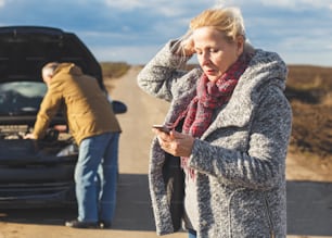 Senior couple on the road having problem with a car, woman calling assistance while man looking at vehicle engine.