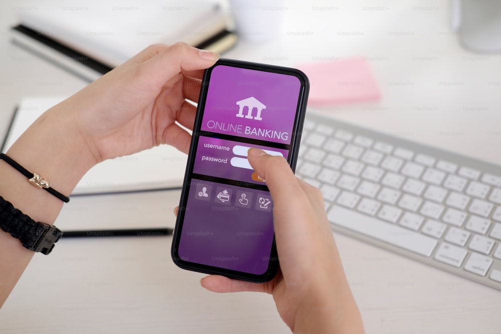 机のワークスペースの画面にオンライン銀行アプリケーションを示すスマートフォンを持つ若い女性