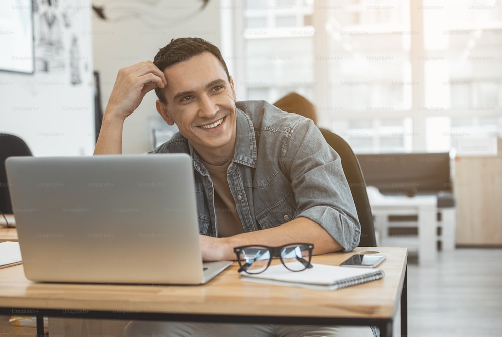 Portrait of cheerful man typing in laptop while sitting at desk in office. Beaming employee at job concept