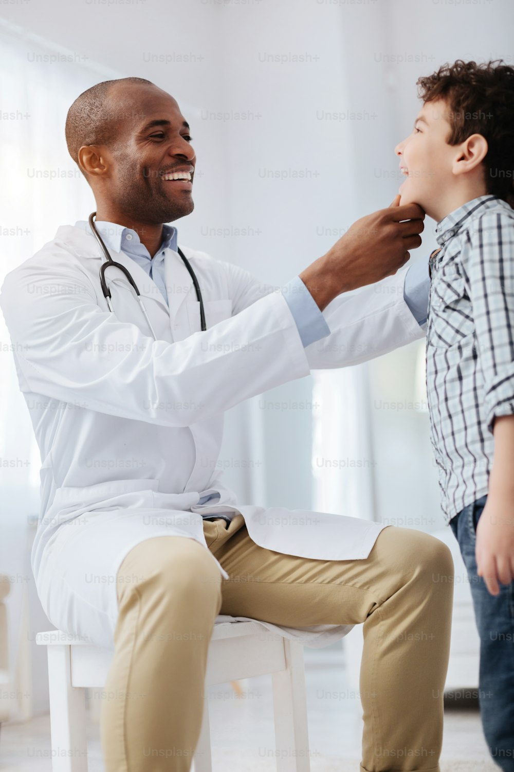 Redness on side. Low angle of professional joyful male doctor looking at boy who staying in profile and checking his throat