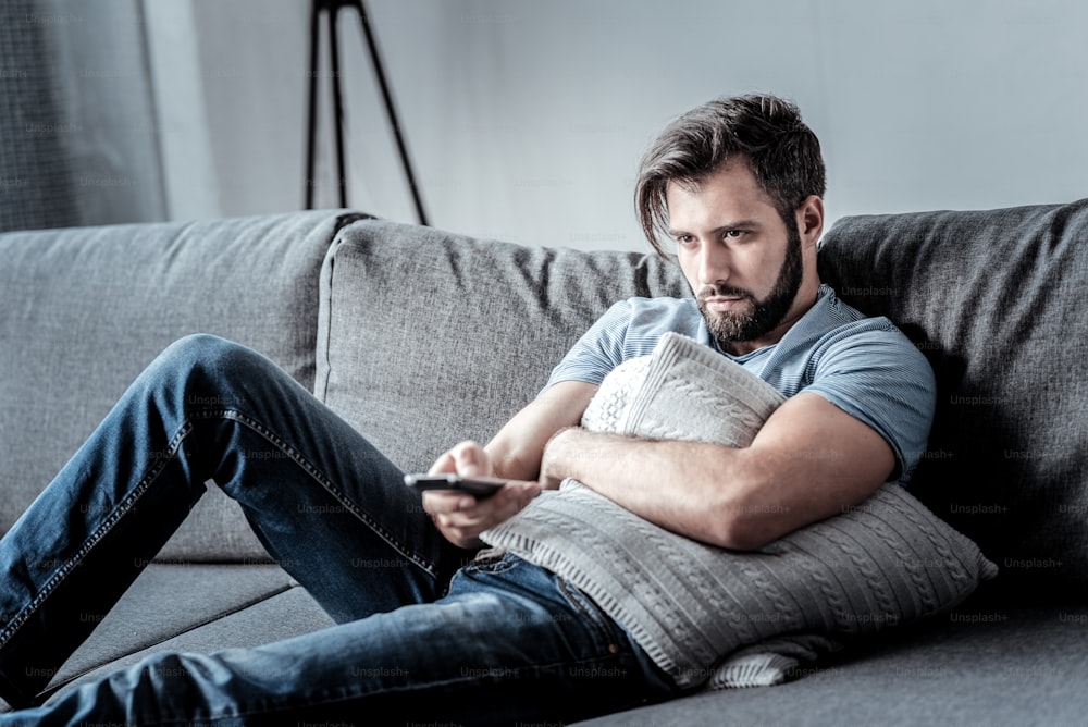 Boring TV shows. Unhappy depressed bored man hugging a cushion and holding a remote control while watching TV shows