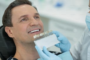 Dientes postizos. Hombre guapo de ojos oscuros mostrando sus dientes y el dentista sosteniendo dientes postizos