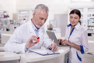 Unir esfuerzos. Médico masculino senior pensativo anotando mientras farmacéutica femenina trabajando en computadora portátil