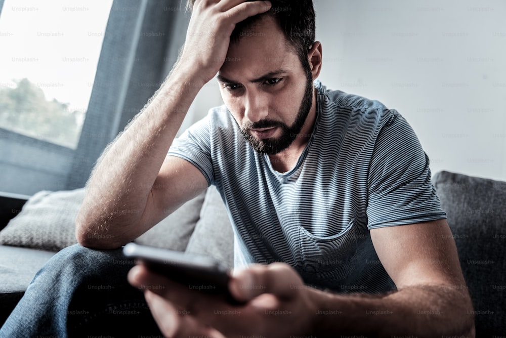 Bad news. Unhappy gloomy cheerless man looking at his smartphone screen and holding his forehead while receiving unpleasant news