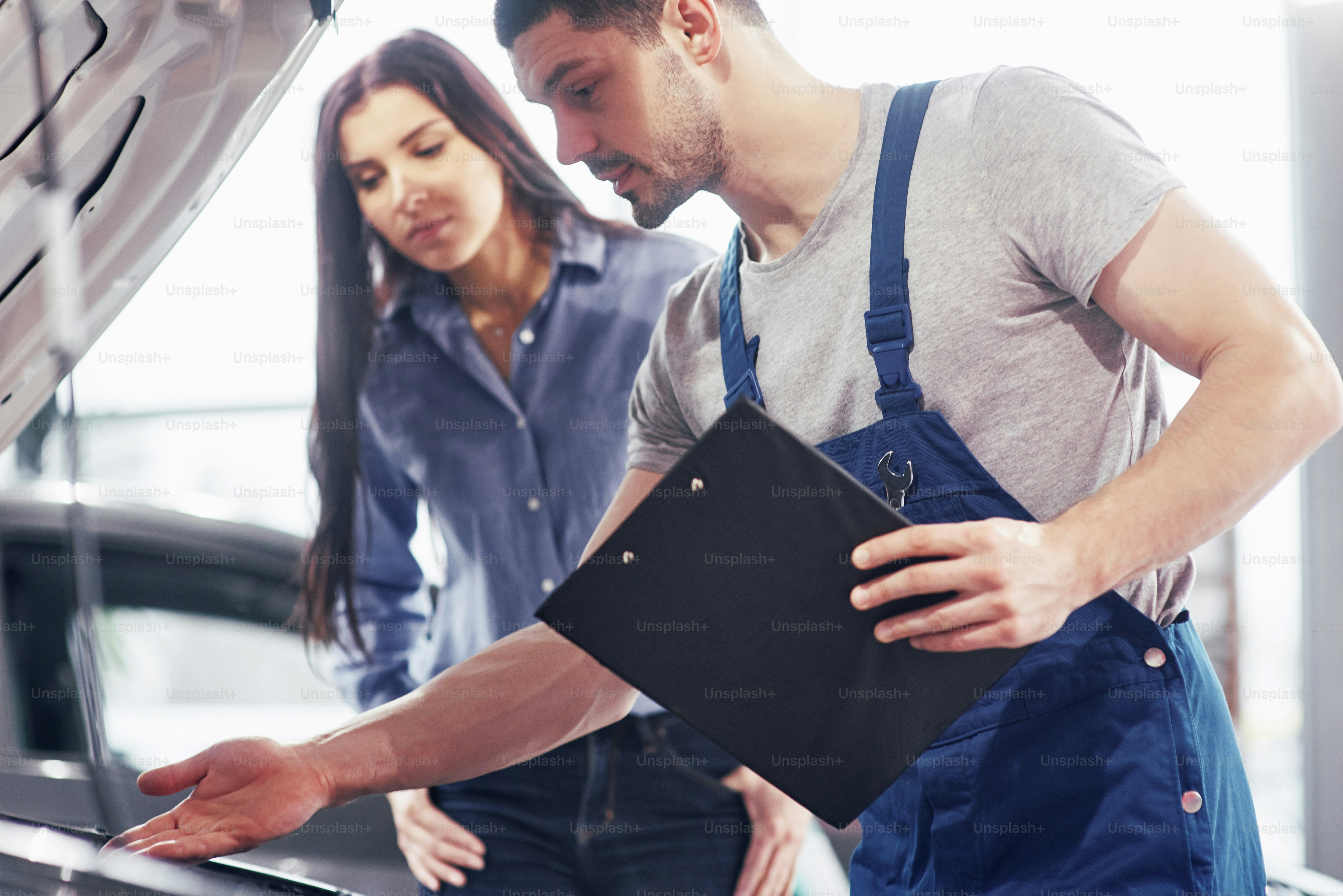 A man mechanic and woman customer look at the car hood and discuss repairs.