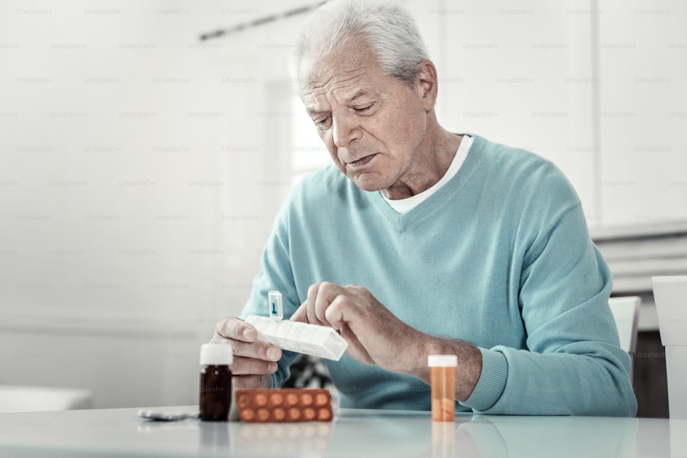 It will helps. Concentrated senior serious man sitting by the table holding pack of tablets and focusing on it.