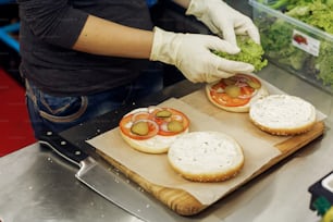 process of making burger. chef hands in gloves cooking hamburgers and cheeseburgers, putting ingridients on wooden desk. catering in food court at mall concept