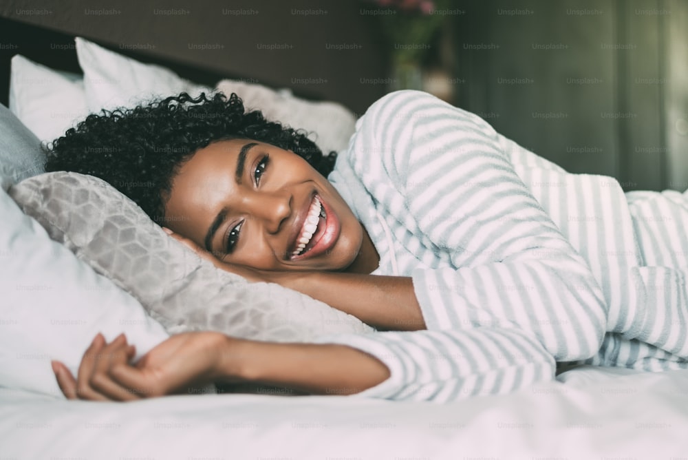 close up of a pretty black woman with curly hair smiling and lying on bed looking at the camera