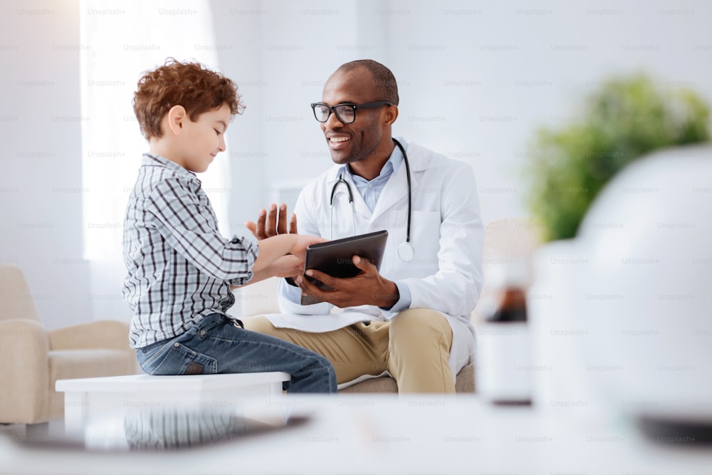 Data in tablet. Energetic cheerful male doctor grinning while showing tablet to boy and sitting