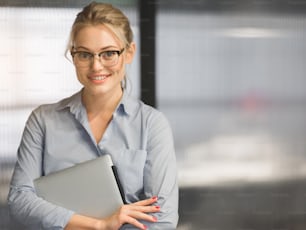 Waist up portrait of confident blond woman holding laptop and smiling. She is standing formalwear and eyeglasses. Profession concept. Copy space