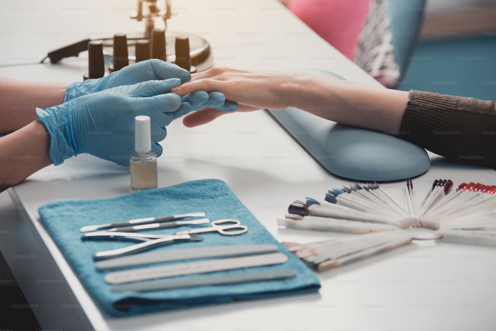 Close up nail artist hand doing manicure procedure for visitor. They situating at desk