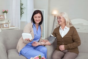 Low blood pressure. Optimistic caregiver using tonometer while elder woman smiling