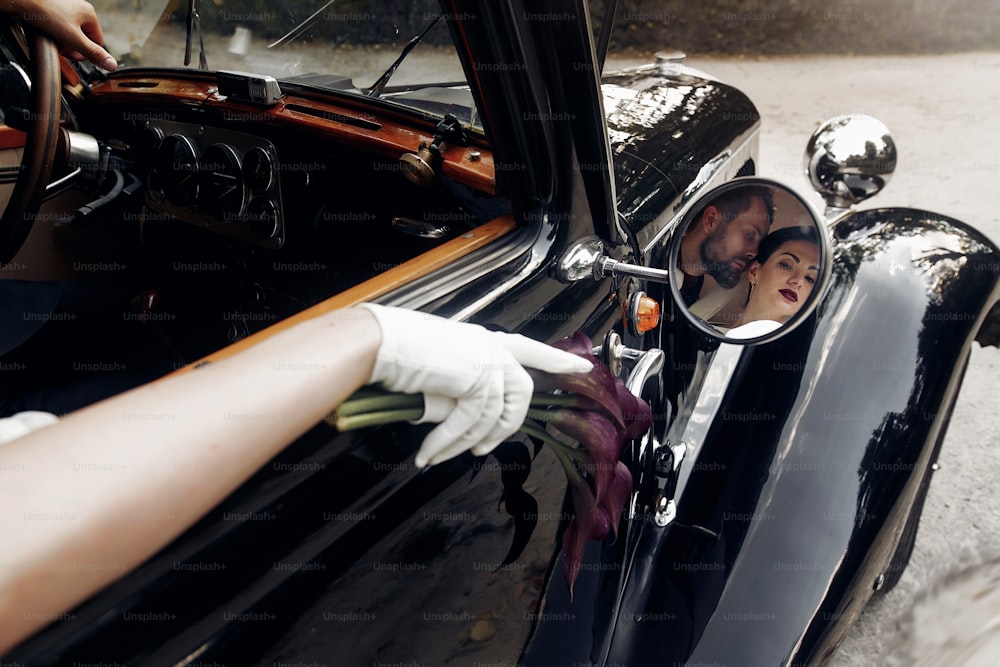 Luxus elegantes Hochzeitspaar, das sich küsst und umarmt in stilvollem schwarzem Auto. ungewöhnlicher Blick im Spiegel. wunderschöne Braut und hübscher Bräutigam im Retro-Stil