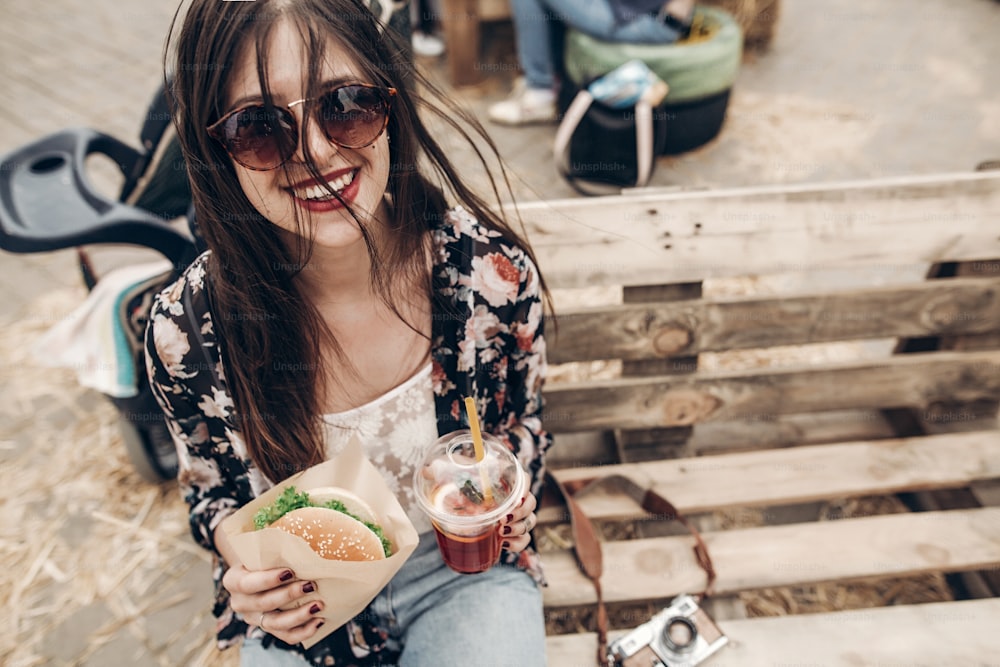 stylish hipster woman holding juicy burger and lemonade. boho girl with hamburger and drink smiling at street food festival. summertime. summer vacation travel. space for text