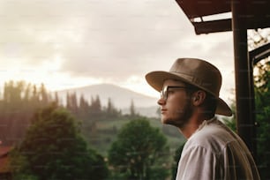 Hipster-Mann auf der Veranda des Holzhauses mit Blick auf die Berge im abendlichen Sonnenuntergang, ruhiger entspannender Moment, Sommerurlaubskonzept, Platz für Text