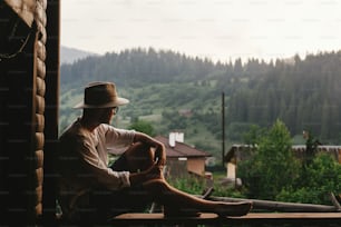 Hipster-Mann sitzt auf der Veranda eines Holzhauses mit Blick auf die Berge am Abend, ruhiger Moment, Sommerurlaubskonzept, Platz für Text