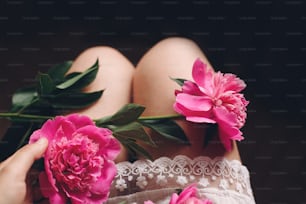 lovely pink peonies on legs of boho girl in white bohemian dress, top view. space for text. stylish hipster woman sitting with beautiful flowers in morning room. atmospheric sensual moment