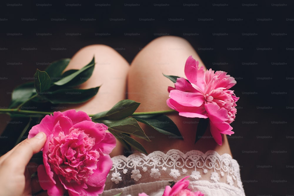 Schöne rosa Pfingstrosen an den Beinen des Boho-Mädchens in weißem böhmischen Kleid, Draufsicht. Platz für Text. Stilvolle Hipster-Frau sitzt mit schönen Blumen im Morgenzimmer. atmosphärisch sinnliches Moment