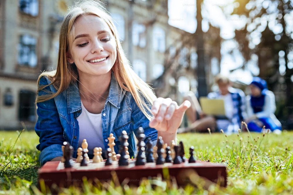 Esto es muy relajante. Hermosa chica rubia sonriendo ampliamente mientras está acostada en la hierba disfrutando del juego de ajedrez al aire libre.