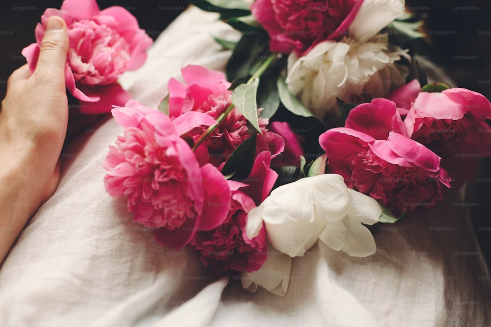 Belles pivoines roses sur les jambes d’une fille bohème en robe bohème blanche, vue de dessus. espace pour le texte. femme hipster élégante assise avec de belles fleurs dans la salle du matin. moment sensuel atmosphérique