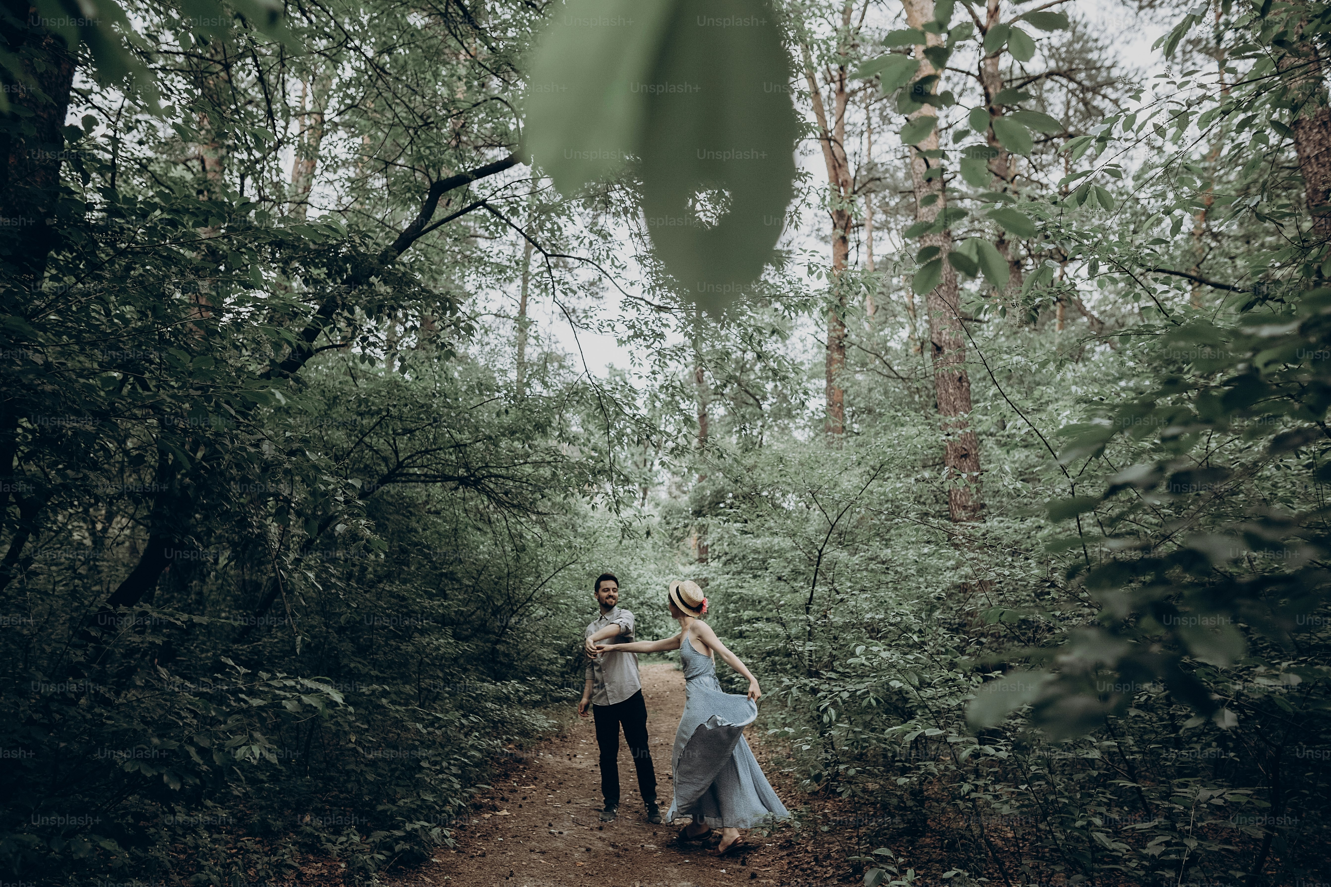 stylish hipster couple walking and dancing in green summer forest. happy man and woman in love, modern outfit, relaxing at park. girl in dress and straw hat with peony. rustic wedding concept