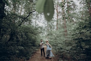 stylish hipster couple walking and dancing in green summer forest. happy man and woman in love, modern outfit, relaxing at park. girl in dress and straw hat with peony. rustic wedding concept
