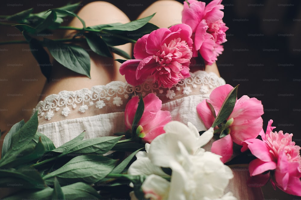 Schöne rosa Pfingstrosen an den Beinen des Boho-Mädchens in weißem böhmischen Kleid, Draufsicht. Platz für Text. Stilvolle Hipster-Frau sitzt mit schönen Blumen im Morgenzimmer. atmosphärisch sinnliches Moment