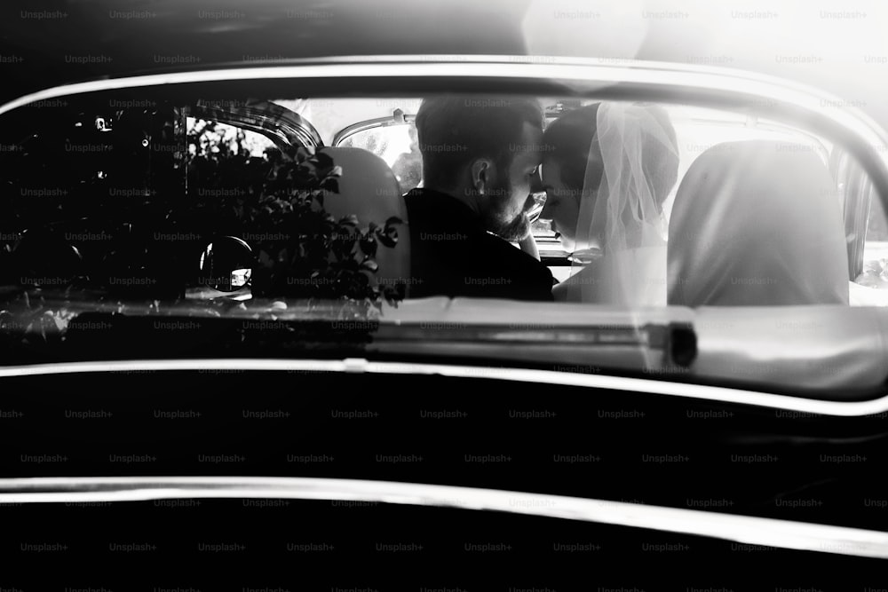 Luxus elegantes Hochzeitspaar, das sich in stilvollem schwarzen Auto in Licht umarmt. Ungewöhnliche Aussicht von hinten. Wunderschöne Braut und hübscher Bräutigam im Retro-Stil. Schwarz-Weiß-Foto