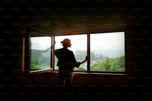 Silhouette der Frau bei Sonnenschein am großen Holzfenster mit Blick auf Sonnenuntergang in den Bergen, ruhiger Moment oder Relax-Konzept, Raum für Text