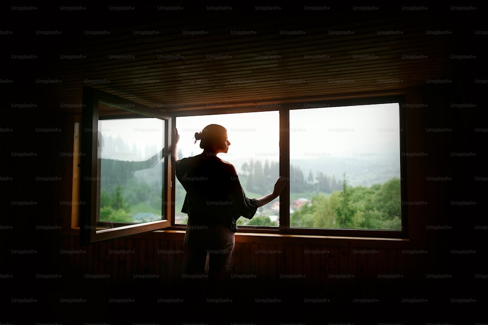 산에서 일몰을 볼 수 있는 큰 나무 창문에서 햇살을 받는 여성의 실루엣, 고요한 순간 또는 휴식 개념, 텍스트를 위한 공간