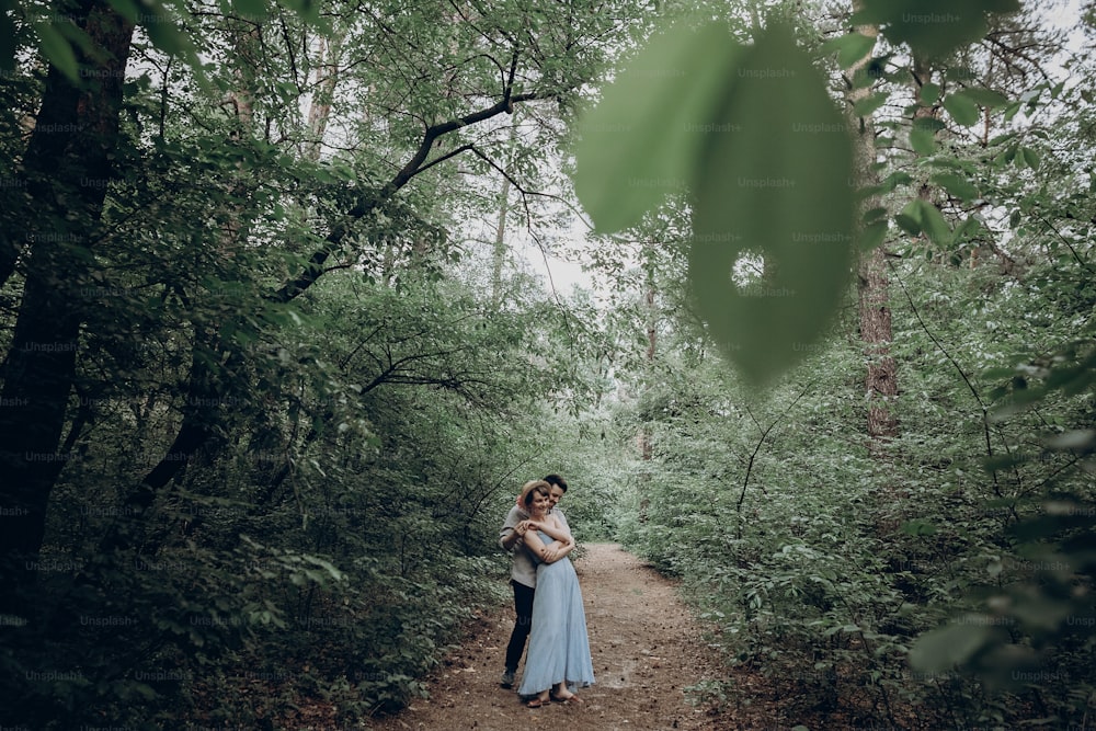세련된 힙스터 신부와 신랑이 녹색 여름 숲에서 걷고 춤을 추고 있다. 사랑에 빠진 행복한 커플, 현대적인 의상, 공원에서 휴식을 취합니다. 드레스와 밀짚 모자를 쓴 소녀. 소박한 결혼식 개념