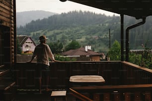 uomo hipster in piedi sul portico della casa di legno e rilassante guardando le montagne in serata, momento calmo, concetto di vacanza estiva, spazio per il testo