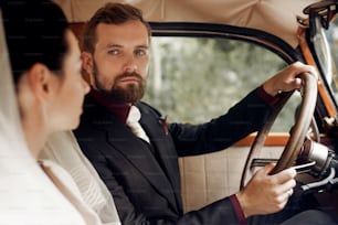 luxury elegant wedding couple posing in stylish black car. handsome groom holding helm  looking at gorgeous bride. retro style. romantic moment