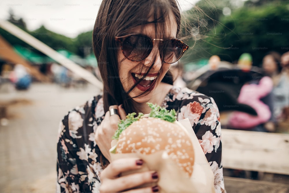 ジューシーなハンバーガーを持って食べるスタイリッシュなヒップスターの女性。ストリートフードフェスティバルで笑顔のハンバーガーを持つ自由奔放に生きる女の子。夏時。夏休み旅行。テキスト用のスペース