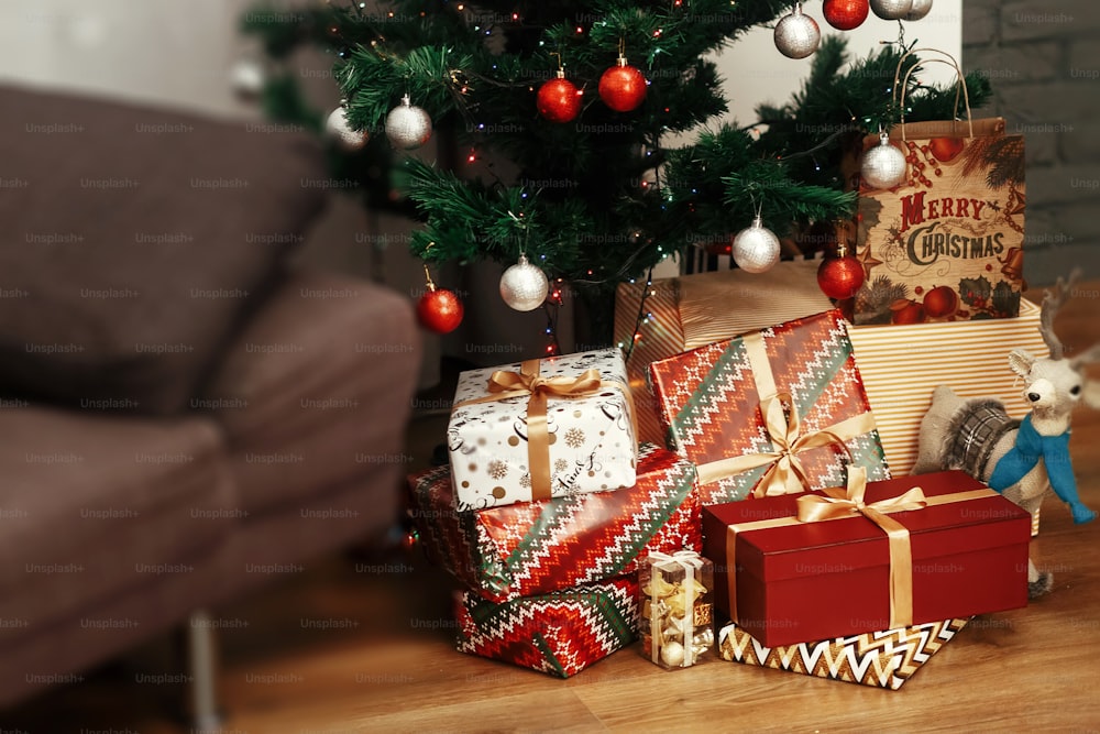 Regali di Natale avvolti in scatole regalo colorate e nastri dorati, sdraiati sullo sfondo del pavimento in legno sotto l'albero di Natale verde con decorazione ornamentale d'argento, concetto di biglietto d'auguri