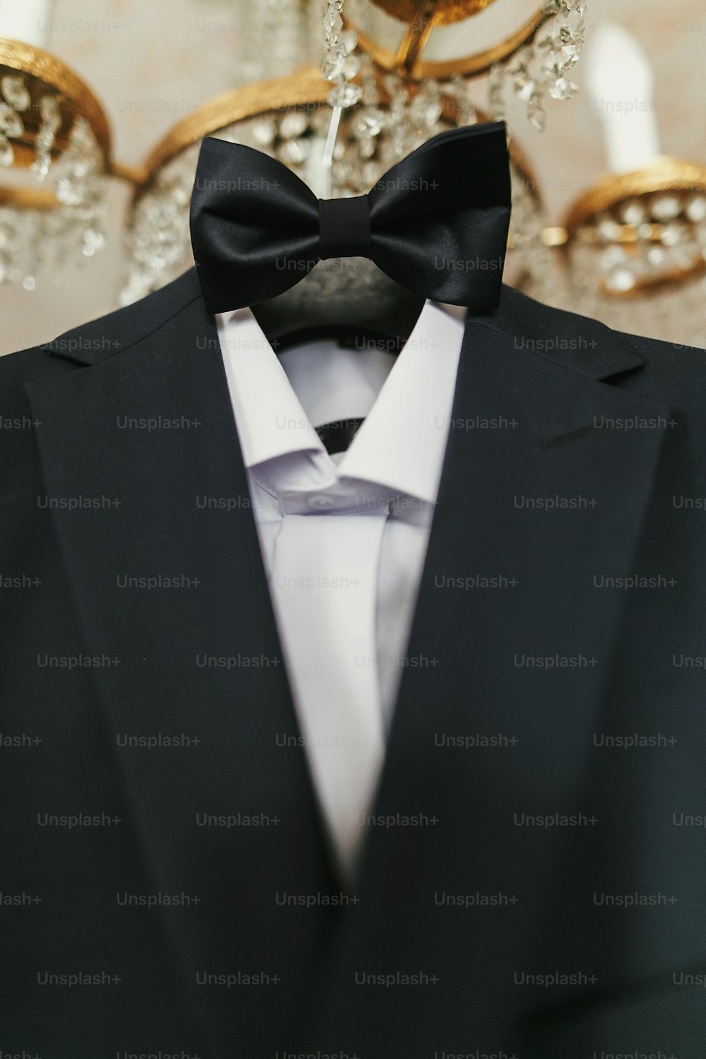 stylish black suit with white shirt and bow tie on hanger on luxury chandelier. morning preparations before wedding day. groom outfit